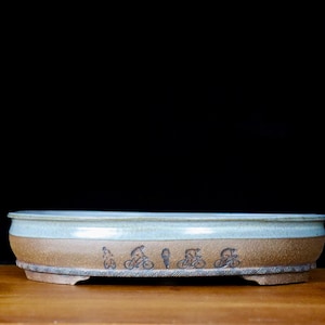 Könnte beinhalten: Ein Keramik-Bonsai-Topf mit hellblauem Rand und braunem Körper. Der Topf zeigt schwarze Fahrradillustrationen um die Mitte. Der Topf steht auf einer Holzoberfläche vor schwarzem Hintergrund.