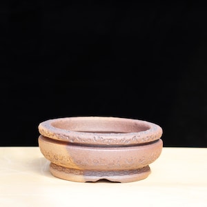 May include: A round, unglazed terracotta bonsai pot. The pot has a wide, shallow bowl shape with a textured rim and decorative patterns around the base. The pot is approximately 20 cm in diameter.