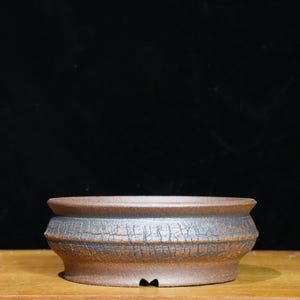May include: A brown, round bonsai pot with a textured, cracked surface. The pot has a wide, shallow design with a slightly raised rim. The pot is made of ceramic and sits on a wooden surface.