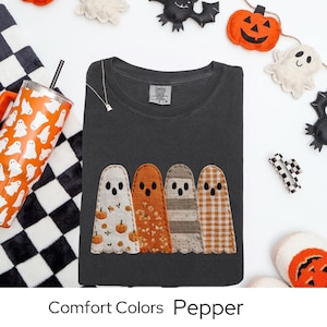 May include: A dark gray Comfort Colors Pepper t-shirt featuring four appliqued ghost figures in various patterns. The ghosts have black dot eyes and are in white, orange, and plaid fabrics. Halloween-themed decorations surround the shirt.