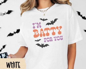 Puede incluir: Camiseta blanca con el texto "I'M BATTY FOR YOU" en morado y naranja, con gráficos de murciélagos negros. La camiseta forma parte de una colección temática de Halloween.