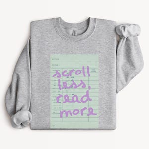 May include: A heather gray crewneck sweatshirt featuring a light green rectangular graphic that resembles a vintage library checkout card. Overlaid on the card is the text "scroll less, read more" written in a casual, handwritten lavender script.