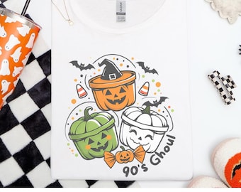May include: White Gildan Softstyle t-shirt with a Halloween design. The graphic shows three cartoon pumpkins in orange, green, and white, with bats, candy corn, and the text "90's Ghoul".