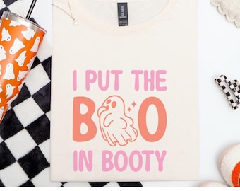 Könnte beinhalten: Ein naturfarbenes Gildan Softstyle T-Shirt mit dem Schriftzug "I PUT THE BOO IN BOOTY" in Rosa und Orange. Eine Geistergrafik befindet sich in der Mitte. Das Shirt hat einen Rundhalsausschnitt.