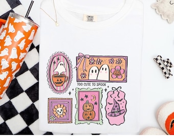 May include: White Comfort Colors t-shirt with a colorful Halloween-themed graphic. The design includes framed illustrations of ghosts, pumpkins, and a witch's hat. The text "TOO CUTE TO SPOOK" is also featured.