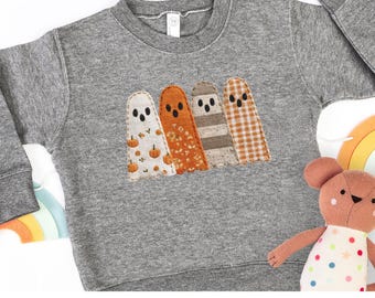 Puede incluir: Una sudadera gris con cuatro figuras de fantasmas aplicadas. Los fantasmas tienen varios diseños, incluyendo calabazas, tela naranja, rayas marrones y un diseño de cuadros. El texto "Rabbit Skins 3317 Granite Heather" está en la parte inferior.
