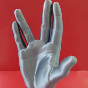 Peut inclure: Sculpture de main de Spock en argent imprimée en 3D avec une base noire. La base porte le texte "LIVE" imprimé en vert et l'insigne de la Starfleet en or.