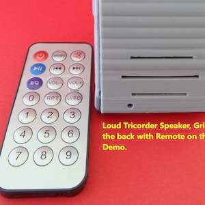 May include: A gray speaker with a grid pattern on the back. A black remote control with a numeric keypad and buttons for play, pause, volume, and mode is included. The text "Loud Tricorder Speaker, Grid on the back with Remote on the left Demo." is printed below the speaker.