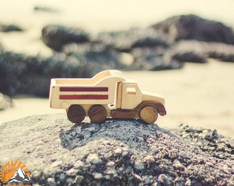 May include: A wooden toy dump truck with a natural wood finish and dark brown wheels. The truck bed features red and brown stripes. The toy is sitting on a rock, with a blurred beach and ocean background.