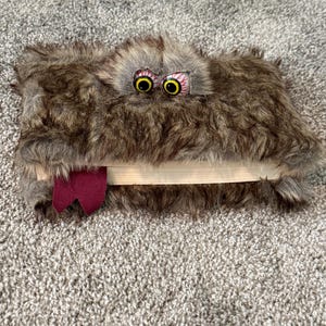 May include: A whimsical, furry book cover with large, googly eyes and a protruding tongue. The cover is a mix of brown and gray faux fur, with a maroon ribbon bookmark. The eyes are yellow and black with red detailing.