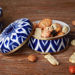 Può includere: Un contenitore in ceramica blu e bianca con un coperchio, riempito con una varietà di noci e snack misti. Il contenitore ha un bordo dorato attorno alla parte superiore.
