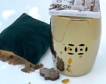 Puede incluir: Un taburete de jardín de cerámica dorada con acabado brillante y patrones decorativos recortados. También hay un cojín de terciopelo verde oscuro con flecos, dos libros y un bloque de madera con un diseño de mano. Las hojas amarillas dispersas añaden un toque otoñal.