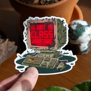 May include: A sticker featuring a vintage computer with the text "AI ART IS CLASS WARFARE" in red. The computer is overgrown with vines and sits on a green base. A mouse and keyboard are also visible.