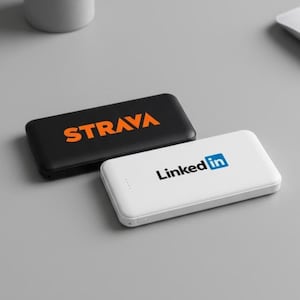 以下が含まれることがあります： 黒地にオレンジ色の「STRAVA」と白地に青色の「LinkedIn」の文字が入った長方形のモバイルバッテリー2つ。グレーの表面に置かれており、背景にはグレーの鉢に入った小さな多肉植物があります。