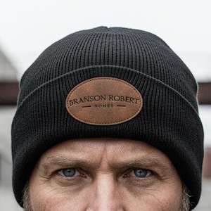Pode incluir: Um gorro preto com um patch de couro castanho com o texto "BRANSON ROBERT - HOME-". A pessoa está usando uma jaqueta jeans preta e tem uma barba completa.