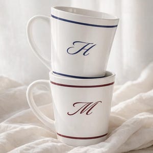 May include: Two white ceramic mugs stacked on top of each other. The top mug has a blue stripe and the letter "H" in a dark blue script. The bottom mug has a burgundy stripe and the letter "M" in a burgundy script. Both mugs have a handle.