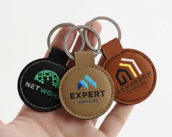 Op de afbeelding: Drie ronde leren sleutelhangers in zwart, beige en bruin, elk met een zilveren sleutelring. De zwarte sleutelhanger heeft een groene afbeelding en het woord "NETWORK". De beige sleutelhanger heeft de tekst "EXPERT SERVICES". De bruine sleutelhanger heeft de tekst "HOMAROOM FURNITURE".