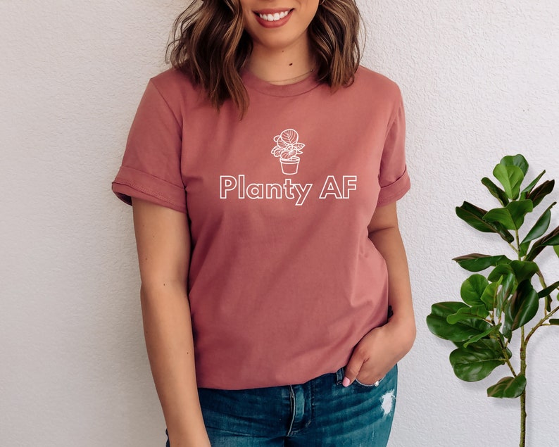 May include: A woman wearing a dusty rose t-shirt with a white outline of a potted plant and the text "Planty AF".