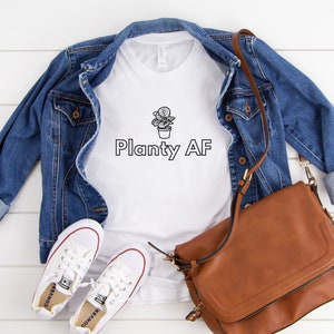 May include: White t-shirt with a black line drawing of a potted plant and the text "Planty AF". The shirt is paired with a denim jacket, a brown leather bag, and white sneakers.