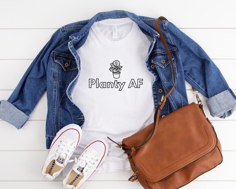 May include: White t-shirt with a black line drawing of a potted plant and the text "Planty AF". The shirt is paired with a denim jacket, a brown leather bag, and white sneakers.
