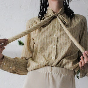 May include: A beige button-up shirt with a small floral pattern and a long tie that is tied into a bow at the neck.