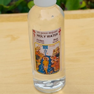 Pode incluir: Uma garrafa de plástico transparente com tampa branca, rotulada como "Holy Water" em inglês e outros idiomas. O rótulo apresenta uma ilustração religiosa. A garrafa contém um líquido transparente.