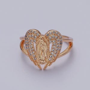 May include: Gold-toned ring featuring a heart-shaped design with a religious figure in the center. The heart is embellished with small, clear stones. The ring has a double-band design.