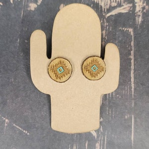May include: A pair of brown and gold earrings with a turquoise square design in the center. The earrings are displayed on a brown cactus-shaped earring stand.