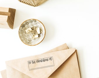 May include: A brown envelope with the text "To the guardian of:" printed on the front. The envelope is on a white surface with a small bowl of gold paper clips and a wooden block.