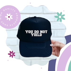 K&ouml;nnte beinhalten: Eine marineblaue Trucker-M&uuml;tze mit dem Text "YOU DO NOT YIELD" in wei&szlig;en Blockbuchstaben. Die M&uuml;tze hat eine Netzr&uuml;ckseite und einen gebogenen Schirm. Ein kreisf&ouml;rmiges Logo mit dem Text "HIGH QUALITY PRODUCTS SHOP" befindet sich in der oberen linken Ecke.