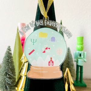 May include: A clear acrylic Christmas ornament shaped like a snow globe with a light blue background. The ornament has a gold ribbon and the text "A few of my favorite things" around the top. Inside the snow globe are various icons including a folder, a psychology symbol, a pencil, a schoolhouse, a brain, a ruler, and a triangle.