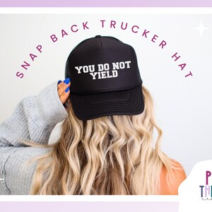 K&ouml;nnte beinhalten: Schwarze Snapback-Trucker-M&uuml;tze mit dem Text "YOU DO NOT YIELD" in Wei&szlig;. Die M&uuml;tze wird von einer Person mit blondem Haar getragen. Die Worte "SNAP BACK TRUCKER HAT" sind &uuml;ber der M&uuml;tze angeordnet. Das Play Therapy Creative-Logo befindet sich unten rechts.