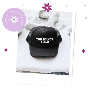 K&ouml;nnte beinhalten: Schwarze Trucker-M&uuml;tze mit Mesh-R&uuml;ckseite und gebogenem Schirm. Auf der M&uuml;tze steht in wei&szlig;er Schrift "YOU DO NOT YIELD". Die M&uuml;tze liegt auf einem wei&szlig;en Kapuzenpullover.