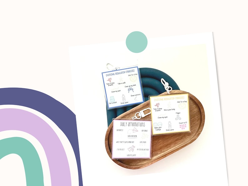 May include: Three small, laminated cards with keychains, featuring text and illustrations for emotional regulation strategies and daily affirmations. The cards are in a wooden tray, with a teal-colored background and a rainbow design in the corner.