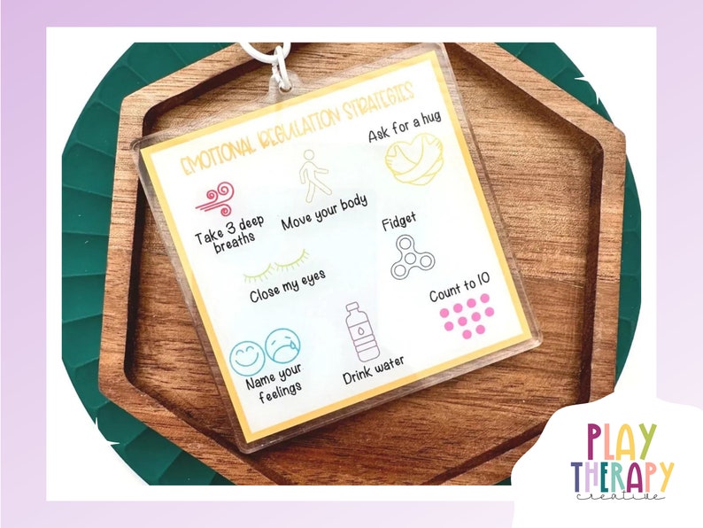 May include: A clear acrylic card with the text "EMOTIONAL REGULATION STRATEGIES" and illustrations of coping mechanisms. The card is on a wooden tray with a green border. The bottom right corner has the text "PLAY THERAPY creative".