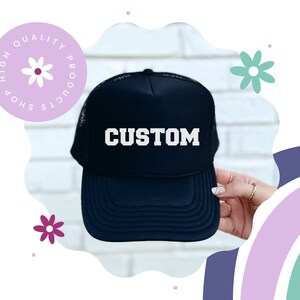 Może przedstawiać: Granatowa czapka typu trucker z napisem "CUSTOM" w białych literach. Czapka ma siatkową tylną część i zakrzywiony daszek. Obraz zawiera dekoracyjny element kwiatowy i tekst "HIGH QUALITY PRODUCTS SHOP".