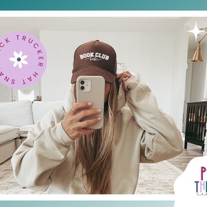 Peut inclure: Un chapeau de camionneur marron avec le texte "Book Club Babe" en blanc. Le chapeau est port&eacute; par une personne aux longs cheveux blonds. La personne prend un selfie dans une pi&egrave;ce avec un canap&eacute; blanc et une biblioth&egrave;que en bois.