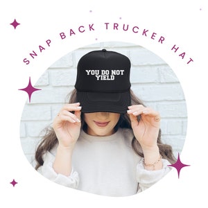 K&ouml;nnte beinhalten: Schwarze Snapback-Trucker-M&uuml;tze mit dem wei&szlig;en Text "YOU DO NOT YIELD". Die M&uuml;tze wird von einer Person getragen. Das Bild ist vor einem wei&szlig;en Hintergrund mit dekorativen Sternformen platziert.