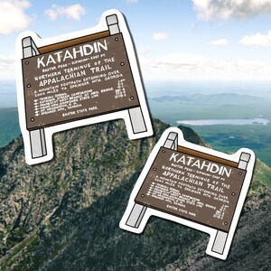 Two stickers of brown wooden sign that sits on peak of Mount Katahdin in Maine. White lettering on sign.  Sticker outlined by white border. Background is real photo from mountain's peaks under cloudy blue sky.