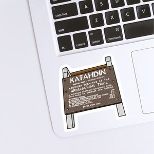 Bottom left corner of silver laptop keyboard. Sticker of brown wooden sign that sits on peak of Mount Katahdin in Maine. White lettering on sign. Sticker outlined by white border.