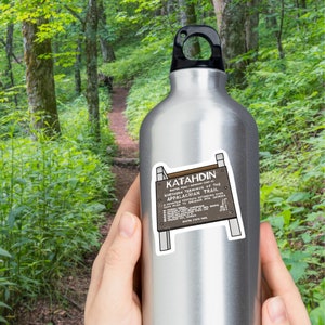 Two hands holding silver canteen with sticker of brown wooden sign that sits on peak of Mount Katahdin in Maine. White lettering on sign.  Sticker outlined by white border. Background is hiking trail with lots of bright greenery.