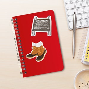 Desktop with red spiral notebook with two stickers on cover. One is brown wooden sign that sits on peak of mount Katahdin in Maine. White lettering on sign. Other is pair of brown leather and rubber-toed boots with yellow laces and white socks.