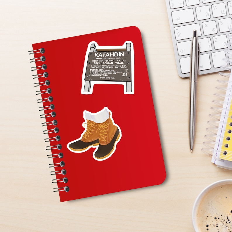 Desktop with red spiral notebook with two stickers on cover. One is brown wooden sign that sits on peak of mount Katahdin in Maine. White lettering on sign. Other is pair of brown leather and rubber-toed boots with yellow laces and white socks.