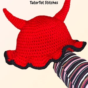 May include: Red crocheted devil hat with black trim. The hat has two horns and a scalloped brim. The hat is on a hand wearing a striped shirt.