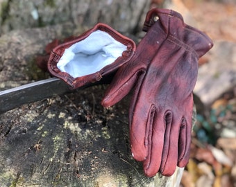 May include: A pair of brown leather gloves with a white interior lining. The gloves are resting on a tree stump with a metal blade visible. The leather has a distressed appearance.