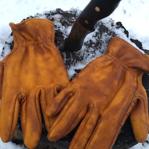 May include: Two pairs of brown leather work gloves on a wooden surface covered in snow. The gloves are worn and have a distressed look.