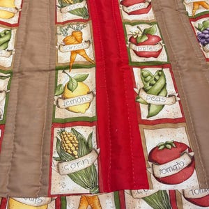 May include: A quilted fabric runner featuring a repeating pattern of various fruits and vegetables including pears, peas, carrots, lemons, corn, apples, grapes, and tomatoes, each labeled with its name on a scroll-like banner.