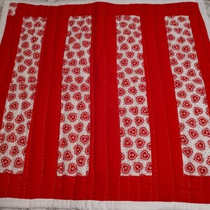 May include: Red and white quilted throw pillow with a heart pattern. The pillow has four red stripes with white stitching.