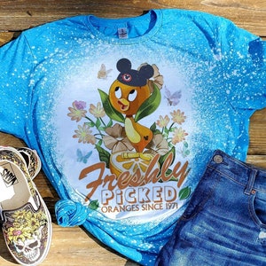 May include: A blue t-shirt with a bleach-splattered design featuring a cartoon duck wearing a Mickey Mouse hat and holding an orange. The text "Freshly Picked Oranges Since 1971" is printed below the duck.