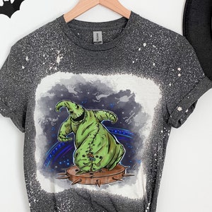 Op de afbeelding: Een grijs T-shirt met een bleekmiddel-gespatte ontwerp met een groen personage uit The Nightmare Before Christmas. Het personage staat op een platform met spikes.